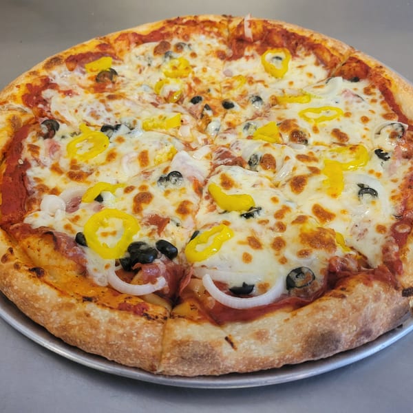 A Free Wheeler Pizza with melted cheese, black olives, onions, and yellow peppers on a metal tray.
