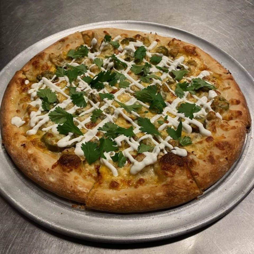 A Free Wheeler Pizza with green chiles, cilantro, and a white sauce drizzle on a metal tray.