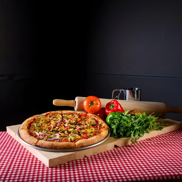 Free Wheeler Pizza with fresh vegetables, rolling pin, and measuring cup on a red checkered tablecloth.