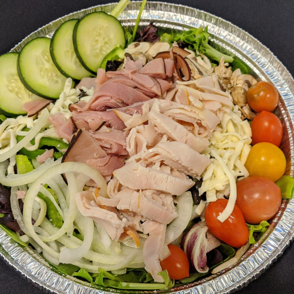 A large salad in a foil pan with ham, turkey, cheese, cucumbers, onions, and tomatoes.